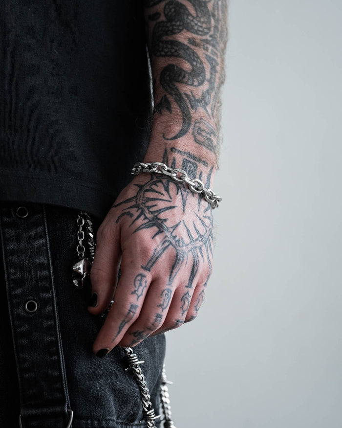 A close-up of a tattooed hand and forearm wearing the 10mm Cable Bracelet. The person has black nail polish, black denim pants with a chain, and visible tattoos. The background is neutral gray.