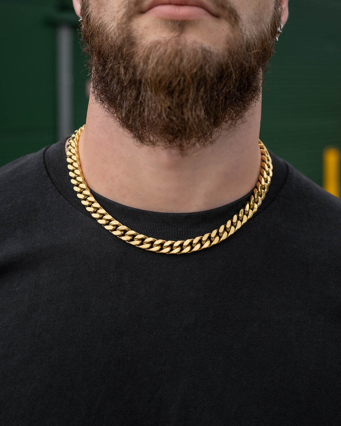 A close-up of a person with a brown beard wearing a black shirt and two thick 11mm Cuban Chain (Gold) necklaces, standing outdoors with a blurred background.