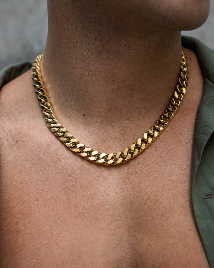 A close-up of a person's neck wearing the 11mm Cuban Chain (Gold), a thick, gold plated necklace styled with an open-collared olive-green shirt for a striking statement look.