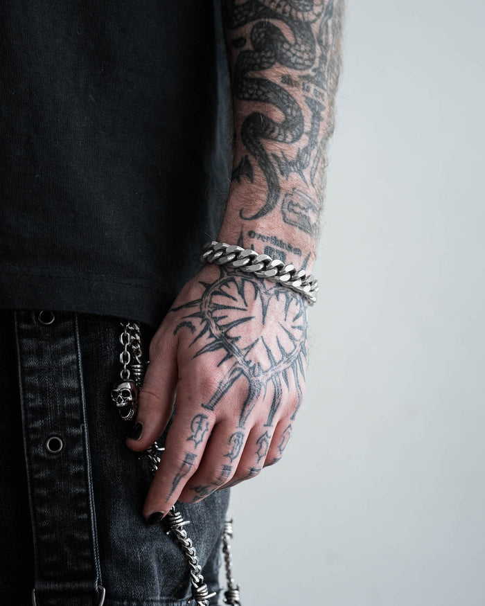A tattooed arm and hand wear the 13mm Chunky Cuban Bracelet in a punk style. The hand features a large clock tattoo and other designs. Black jeans with metal chains and a black shirt complete the look against a light, plain background.