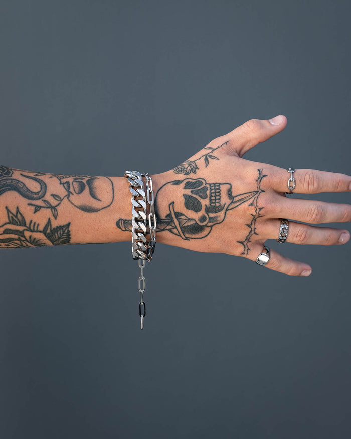 A tattooed hand wearing silver rings and the 13mm Cuban Cluster Bracelet with a box lock is shown against a plain gray background. The tattoos feature a skull, flowers, and patterns, with fingers slightly spread.