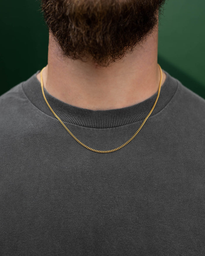 A close-up of a person in a gray crew-neck shirt wearing the 2.3mm Curb Chain (Gold) necklace, featuring a thin gold plated stainless steel design, with a short brown beard and a green background.