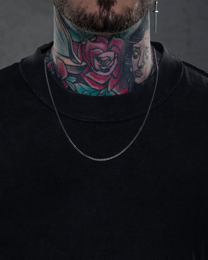 A person wearing a plain black shirt, a 2.3mm Curb Chain necklace, and a cross earring in their left ear. Their neck is covered in colorful tattoos of roses and a woman's face.