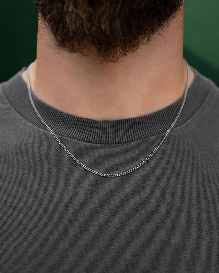 A close-up shows a person wearing Statement Collective's 2.3mm Curb Chain over a dark gray crew neck shirt, with only their neck and upper chest visible. A softly blurred green background highlights the chain's elegance.