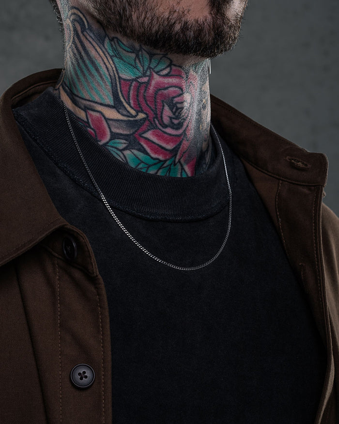 Wearing a brown jacket and black shirt, a person with a colorful rose-and-leaf neck tattoo accessorizes with a 2.3mm Curb Chain necklace. The photo focuses on the neck, clothing, and necklace, with the face partially visible.