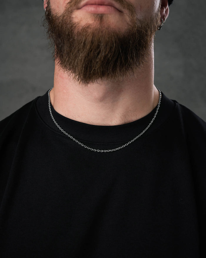A close-up of a man in a black shirt wearing the 3mm Cable Chain, a thin, non-tarnish stainless steel necklace resting just above his collarbone. He has a full brown beard and stands against a plain dark gray background, face partially out of frame.