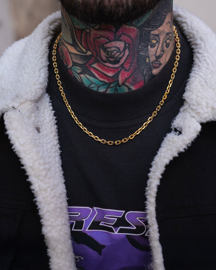 A close-up of a person wearing the Statement Collective 4.5mm Cable Chain (Gold), a black shirt with purple text, and a sherpa-lined jacket. Their neck and lower face feature tattoos of a rose and a woman's face.