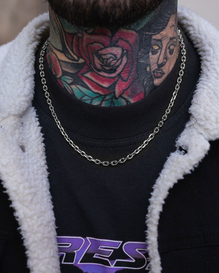 A close-up of a person's tattooed neck, featuring a rose and woman's face, wearing Statement Collective's 4.5mm Cable Chain necklace, a black T-shirt with purple lettering, and a black jacket with a white fleece collar.