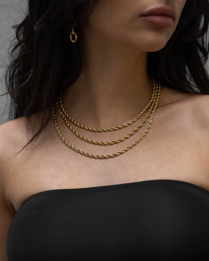 A woman in a strapless black top wears layered gold necklaces and a gold hoop earring. Her dark, wavy hair partly covers her face, drawing attention to the elegance of the 4mm Rope Chain (Gold) around her neck.