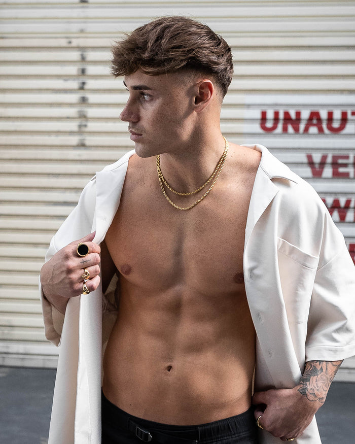 A shirtless man with short brown hair stands in front of a metal shutter, wearing the 4mm Rope Chain (Gold), rings, and a white open shirt draped over his shoulders as he looks to his left.