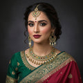 An ornate Indian traditional jewelry set featuring a gold-plated necklace, matching earrings, and a maang tikka, embellished with white faux pearls, gold-toned metal, and multicolored stones, displayed on a dark reflective surface with the brand logo 'SAHELI MY BEST FRIEND' in the background.