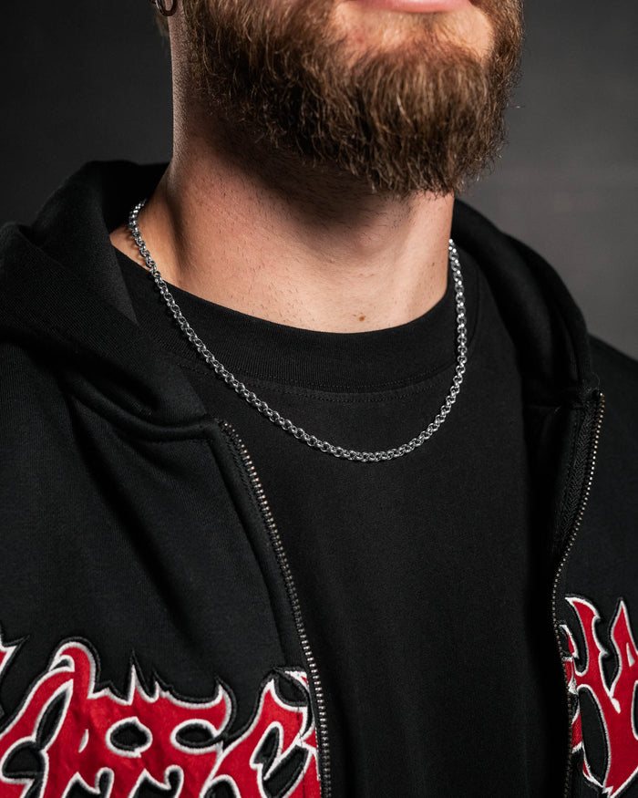 A bearded person wearing a 5mm Rolo Chain necklace, a black t-shirt, and a black zip-up jacket with red and white flame embroidery, shown from shoulders to chin against a dark background.