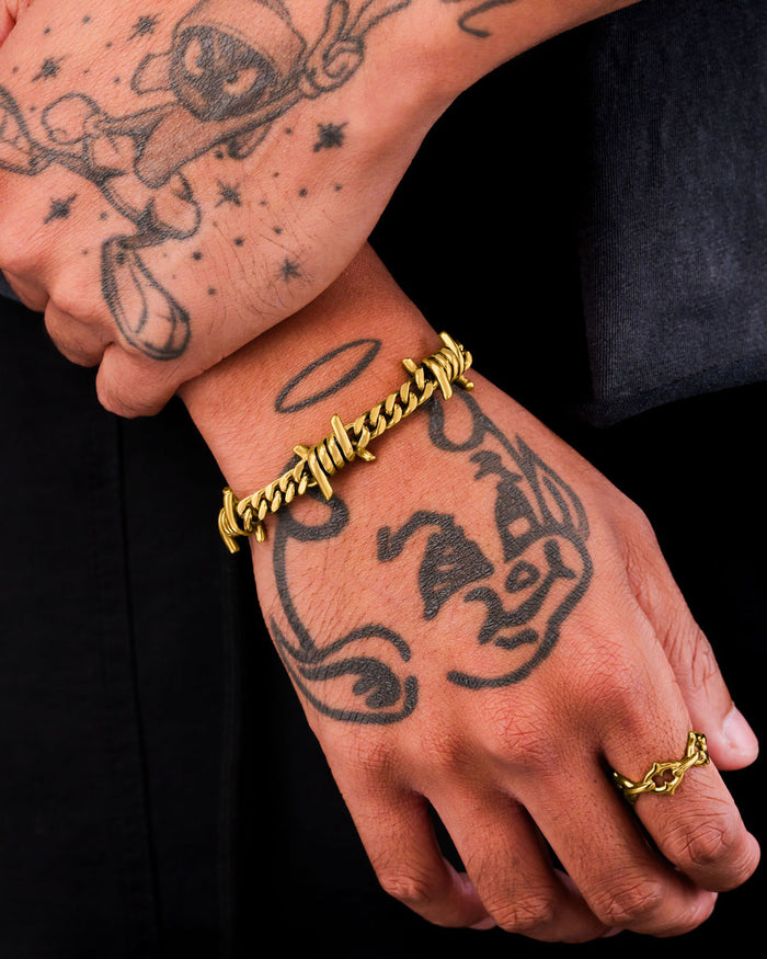 A close-up of tattooed hands wearing the 6mm Barbed Wire Cuban Bracelet (Gold) and a matching gold ring, featuring cartoon tattoos with a halo and hat, set against a black background for a bold jewelry statement.