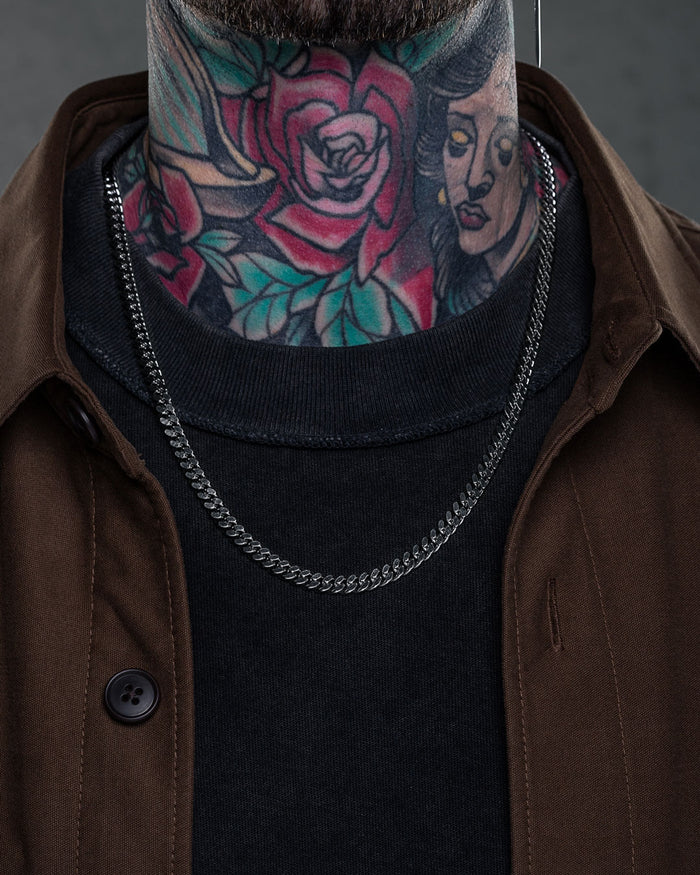 A person in a black shirt, brown jacket, and 6mm Cuban Chain shows off colorful neck tattoos featuring a rose and a woman's face.