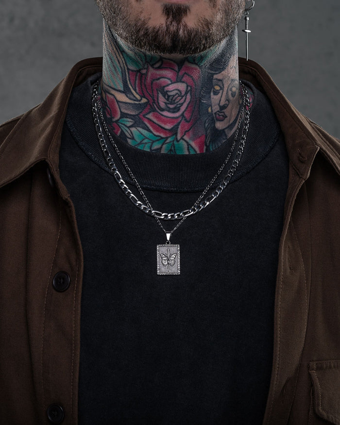 A person with colorful neck tattoos wears layered silver chains, including the 6mm Figaro Chain with a square pendant featuring a leaf design, along with a black shirt and brown jacket. Their face is partially visible.