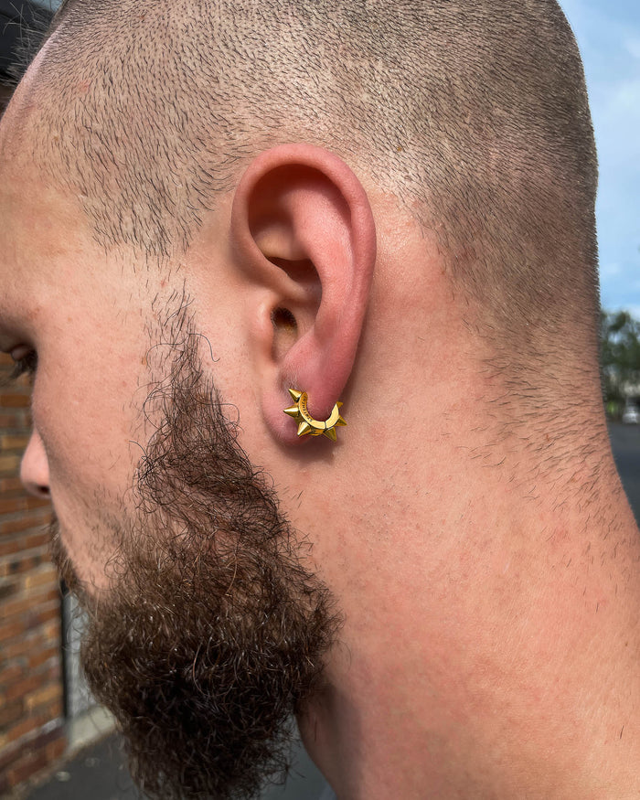 Close-up of a person's ear wearing 6mm Spiked Hoop Earrings (Gold) with a trimmed beard and short hair. Set against an outdoor brick wall and trees, these hypoallergenic hoops add an edgy touch to any look.