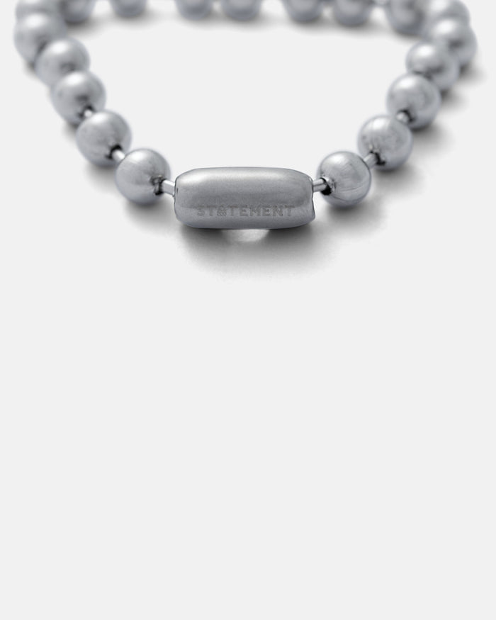 A close-up of the 7mm Ball Bracelet, a silver ball chain with a rectangular STATEMENT-engraved clasp, inspired by vintage 90s style, displayed on a plain grey background.