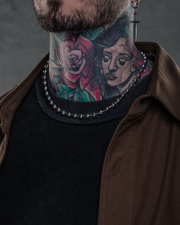 Wearing a dark shirt, brown jacket, cross earring, and the 7mm Ball Chain necklace, a person with colorful neck tattoos—featuring a rose and woman's face—is shown with only the lower half of their face visible.