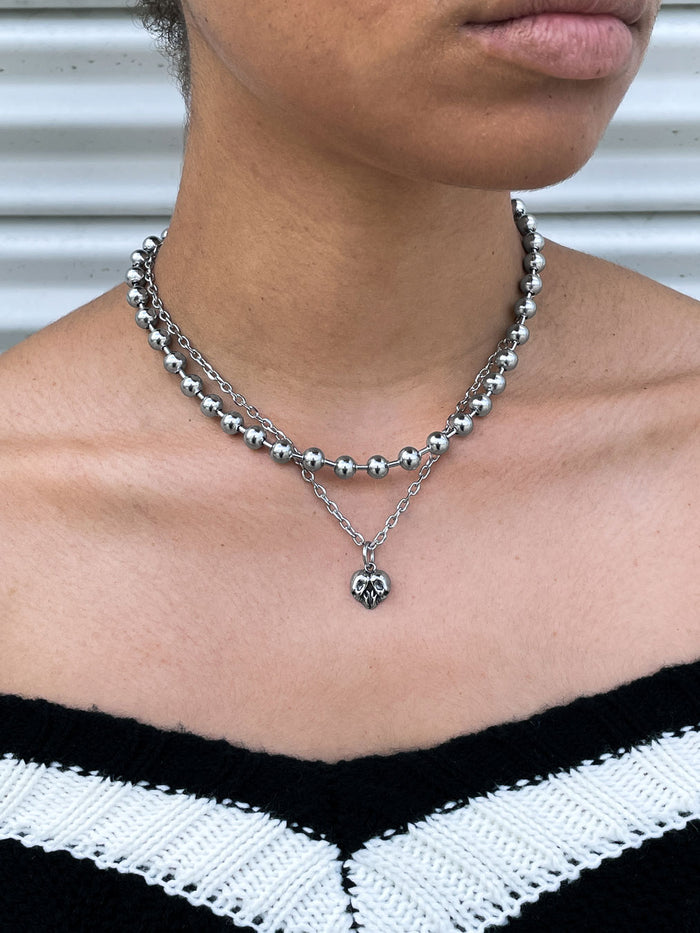A person wearing layered silver necklaces, including a 7mm Ball Chain with large beads and a heart pendant, paired with a black and white off-the-shoulder zigzag top—classic mall goth jewelry style.