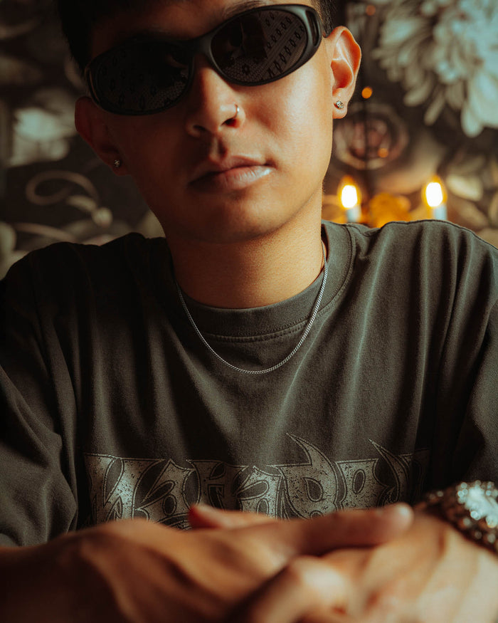 Wearing dark sunglasses, a nose ring, earrings, and a patterned T-shirt, the person sits with folded hands as their 925 Sterling Silver 2.3mm Curb Chain necklace subtly peeks out. Floral decor and glowing candles add to the tranquil ambiance.