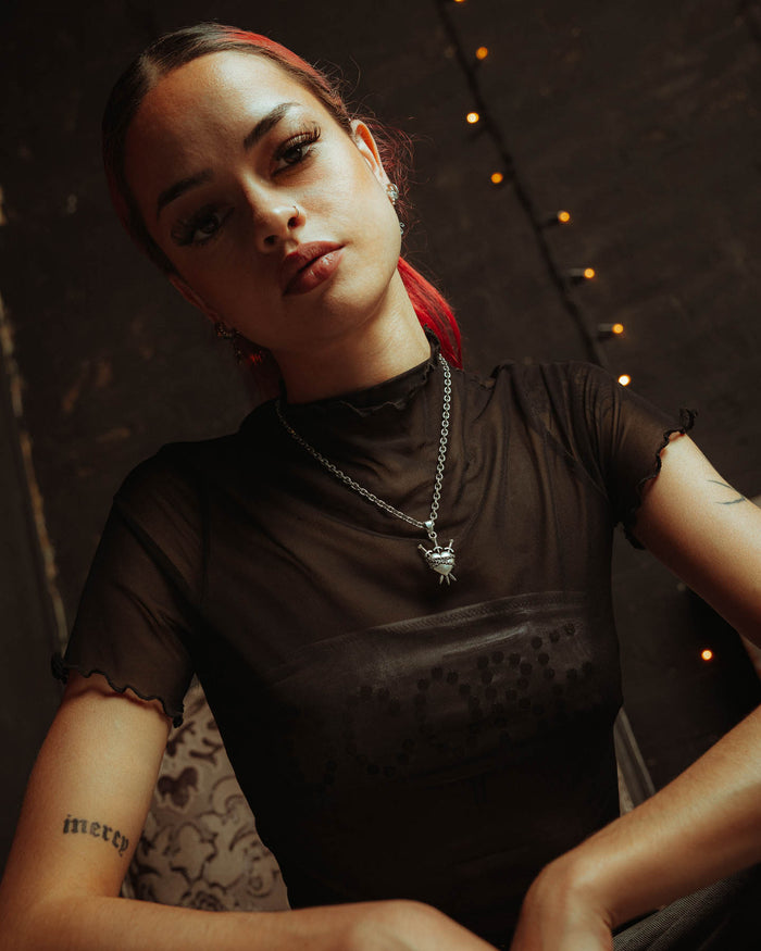 A person with red hair tied back wears a black sheer top, the 925 Sterling Silver "3 of Swords" Pendant necklace, and stud earrings. Tattoos cover their arms as they sit before a dim, string-lit backdrop, gazing confidently at the camera.