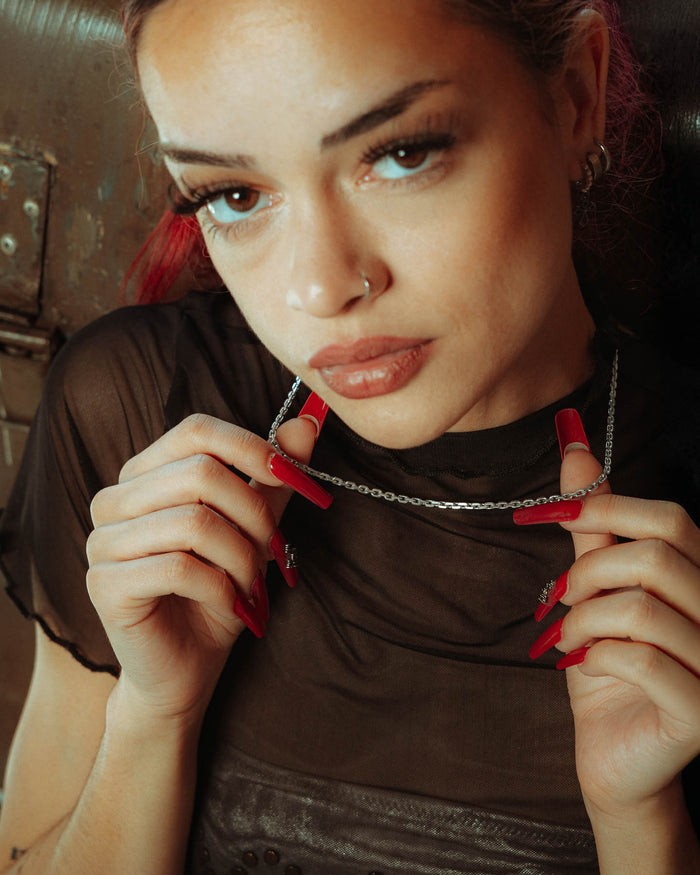 A woman with long red nails and red lipstick holds the 925 Sterling Silver 3mm Cable Chain near her face. She wears a nose ring, hoop earrings, and a sheer black top, looking directly at the camera with a neutral expression.
