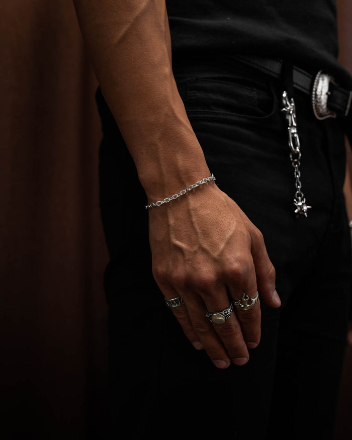 male model in tight black compression top holding first up to show the 4.5mm cable bracelet in sterling silver around his wrist with hand flat against leg to show off flame ring, oval signet ring and roman numerals ring in sterling silver.