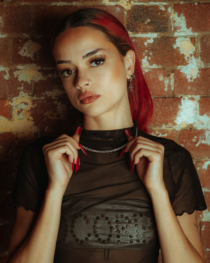 A woman with red hair and bold red nails, wearing a sheer black top, confidently holds a 925 Sterling Silver 4.5mm Cable Chain against a brick wall as she looks at the camera.
