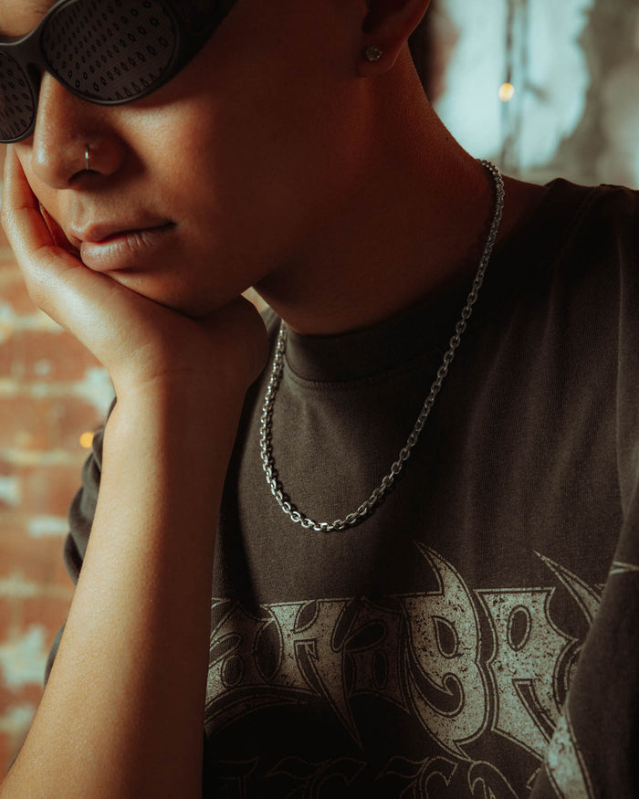 A person wearing dark sunglasses, a nose ring, and the 925 Sterling Silver 4.5mm Cable Chain rests their chin on their hand, sporting a black graphic tee with a brick wall and blurred hanging lights in the background.