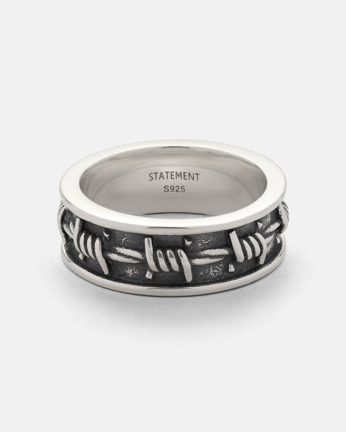 Introducing the 925 Sterling Silver Barbed Wire Ring by Statement Collective. This striking stackable ring features a raised, 3D barbed wire band that wraps around a polished sterling silver edge, set against a dark, textured background for eye-catching contrast. Inside the band, “STATEMENT S925” is finely engraved—a bold accessory with modern style and meticulous detail.
