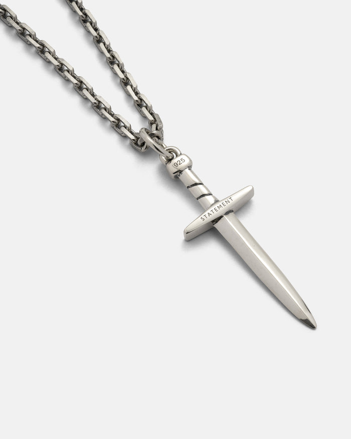 The 925 Sterling Silver Dagger Pendant by Statement Collective is showcased on a light background. This eye-catching necklace features a silver chain and a handmade dagger pendant, with "STATEMENT" engraved on the hilt and "925" on the handle. Expertly crafted from .925 sterling silver, it offers both style and lasting quality.