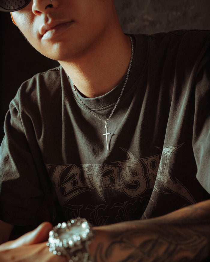 A person wears a dark graphic t-shirt, a cross necklace, and a 925 Sterling Silver Dagger Pendant. Their tattooed arm is visible, and their face is partially out of frame and lit by soft light.