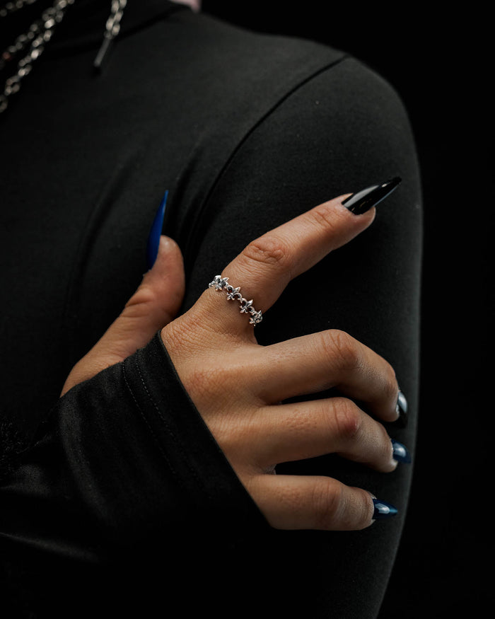A hand with long, pointed black and blue nails wears the Statement Collective 925 Sterling Silver “Fleur De Lis” Ring, its antique silver finish adding a touch of elegance as the person, dressed in a fitted, long-sleeved black top, poses against a dark background.