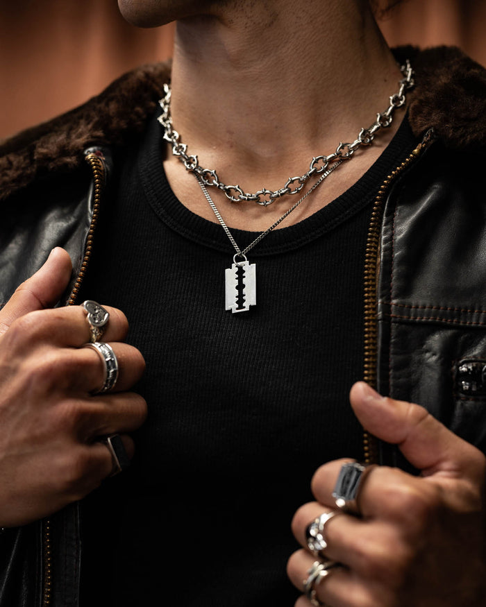 A person in punk metal style—silver rings, black shirt, leather jacket—shows off two necklaces: a chunky chain and the 925 Sterling Silver Razor Blade Pendant, holding their jacket open.