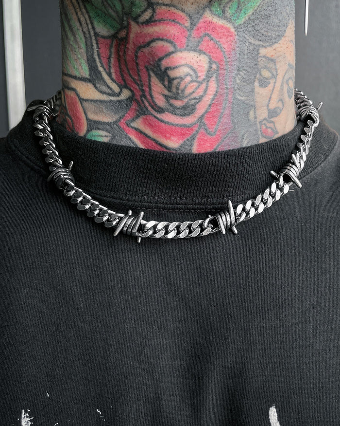 A close-up shows a person wearing the 9mm Barbed Wire Cuban Chain around their neck, featuring colorful rose and woman's face tattoos, paired with a black shirt.