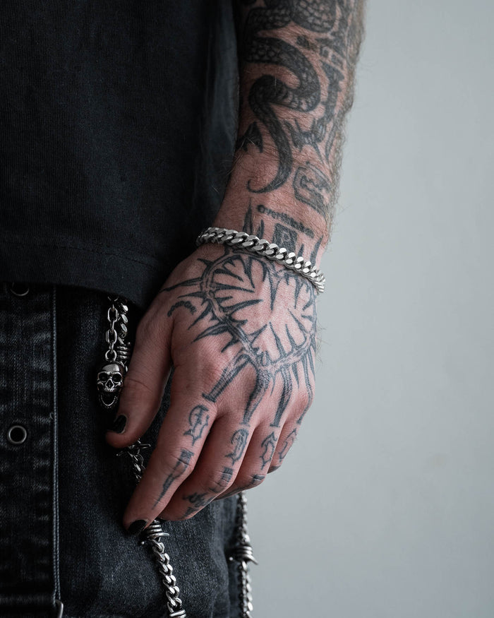 A close-up of a tattooed hand with black nail polish wearing the 9mm Cuban Bracelet. The person is dressed in dark clothing, and a skull chain accessory is attached to their pants.