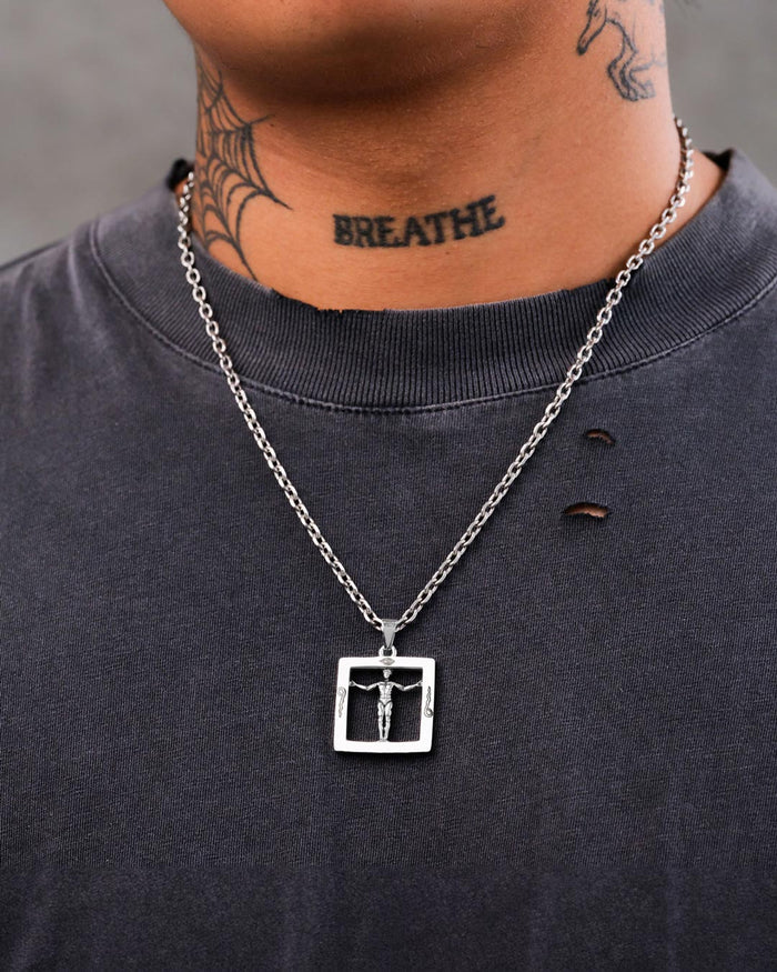 A person sports a dark, distressed t-shirt and the Agrippa’s Square Pendant—a silver occult necklace. They have a BREATHE tattoo and a spiderweb tattoo on their neck.
