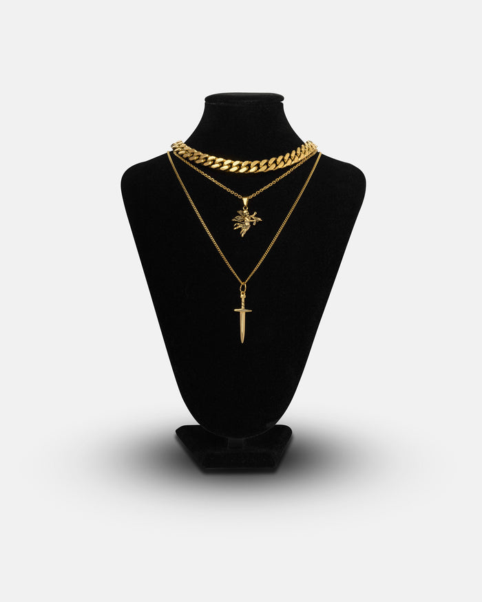 A black jewelry display showcases the Angel & Dagger Set (Gold) by Statement Collective, featuring a striking trio of gold necklaces: a bold Cuban link chain, one necklace with an intricate dragon pendant, and another adorned with a vertical sword charm—all elegantly layered against a plain white background.