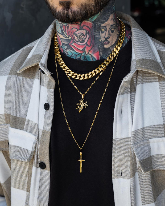 A person wearing the Statement Collective Angel & Dagger Set (Gold)—a striking stack of three gold necklaces, including a bold Cuban link and a pendant—layered over a black shirt and plaid overshirt. Their neck is adorned with colorful tattoos, featuring a vivid red rose and a woman's face.