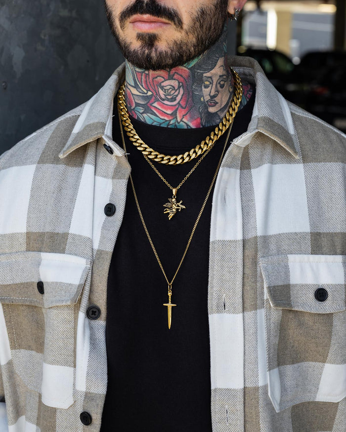 Wearing a black shirt layered under a beige plaid overshirt, a person with neck tattoos showcases the Statement Collective Angel & Dagger Set (Gold)—a bold necklace stack featuring layered gold chains, including a thick Cuban link, an angel pendant, and a sword pendant.