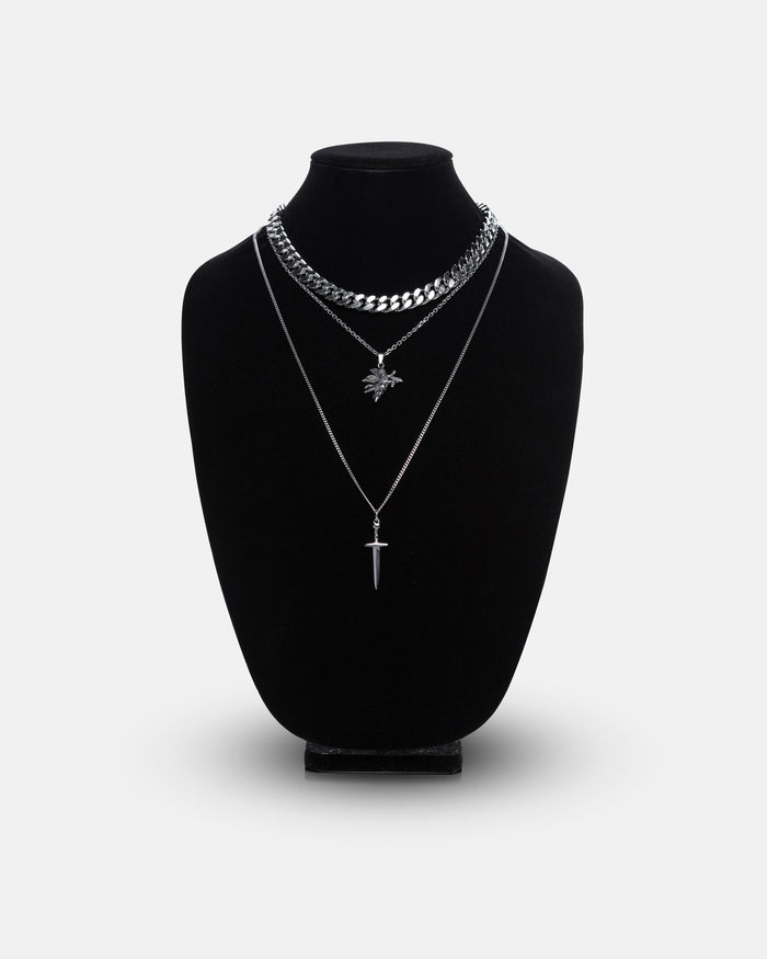 A black velvet necklace bust displays the Statement Collective Angel & Dagger Set: three layered silver necklaces including a bold Cuban link, a delicate chain with a leaf pendant, and another with a dagger pendant, all set against a plain white background.