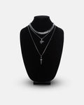 A black velvet necklace bust displays the Statement Collective Angel & Dagger Set: three layered silver necklaces including a bold Cuban link, a delicate chain with a leaf pendant, and another with a dagger pendant, all set against a plain white background.