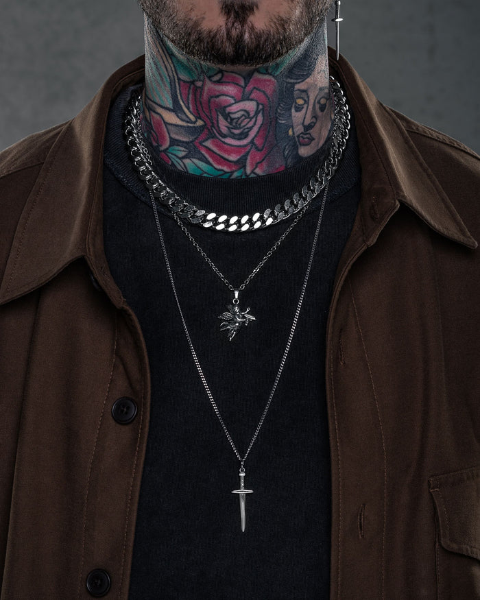 A person with colorful neck tattoos wears layered silver chain necklaces, featuring the Statement Collective Angel & Dagger Set and a classic pendant necklace, over a black shirt and brown jacket.