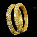 Gold bangles with embedded stones on a dark background, featuring the brand 'Saheli'.