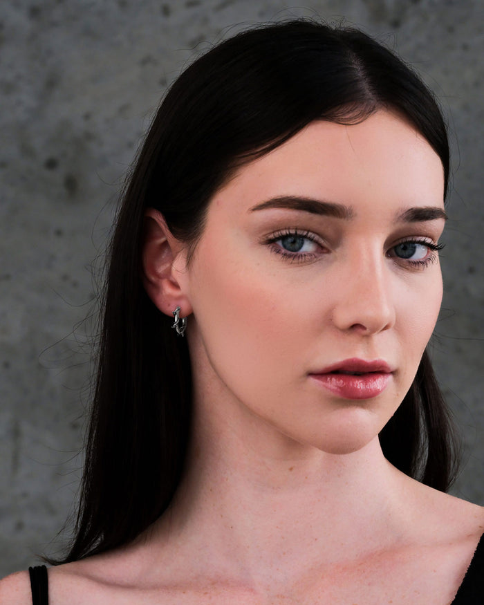 A woman with long dark hair and light skin wears Barbed Wire Hoop Earrings and a black top, looking at the camera with a neutral expression. The background is a plain, gray textured wall.