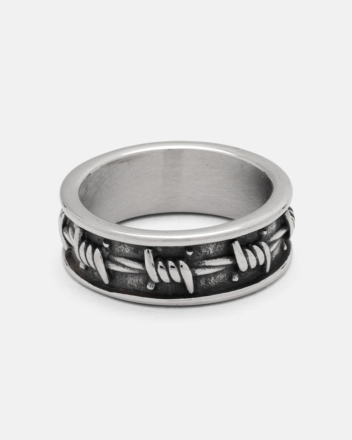 The Barbed Wire Ring by Statement Collective features a silver, twisted barbed wire design crafted from durable 316L stainless steel, with textured detailing and a darkened background for striking contrast, displayed on a plain white surface.