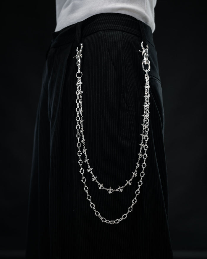 Close-up of a person wearing black pants with the Statement Collective “Baroque” Fleur De Lis Wallet Chain—two sterling silver decorative chains attached to the waistband, hanging in loops on the side like a stylish accessory—set against a dark background.