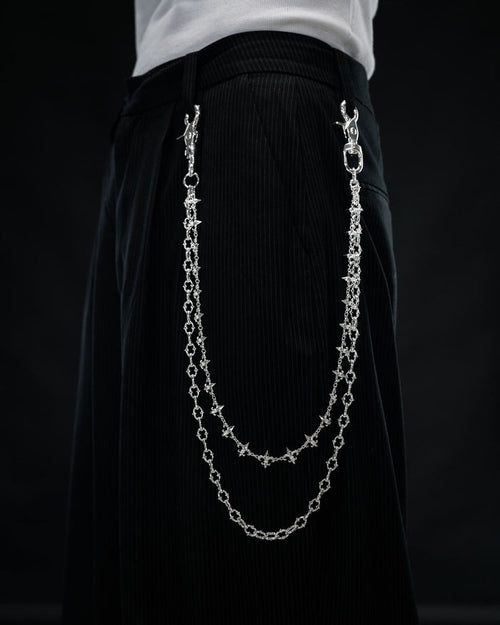 Close-up of a person wearing black pants with the Statement Collective “Baroque” Fleur De Lis Wallet Chain—two sterling silver decorative chains attached to the waistband, hanging in loops on the side like a stylish accessory—set against a dark background.