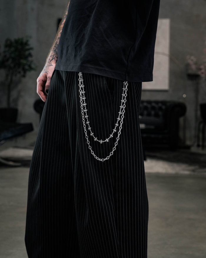 A person wearing black pinstripe pants accessorized with the “Baroque” Fleur De Lis Wallet Chain by Statement Collective, paired with a black t-shirt. The background features a dark sofa and a blurred indoor setting.