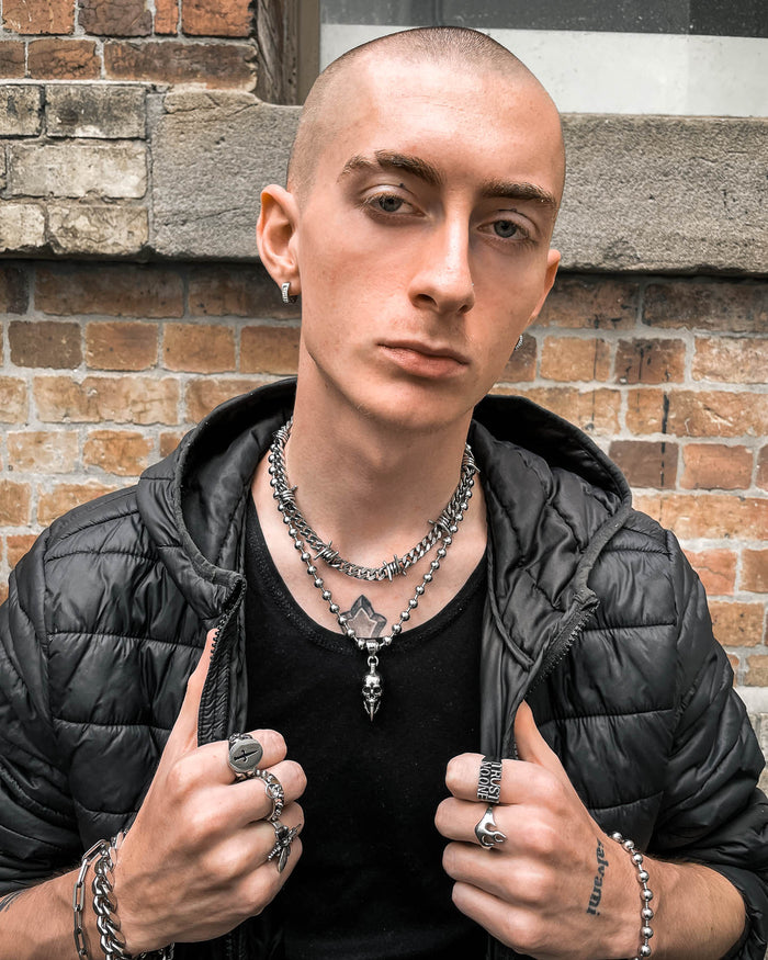 A person with a shaved head and eyebrow piercing wears layered silver necklaces featuring the "Bite The Bullet" Pendant, along with rings, hoop earrings, a black shirt, and jacket, standing confidently before a brick wall.